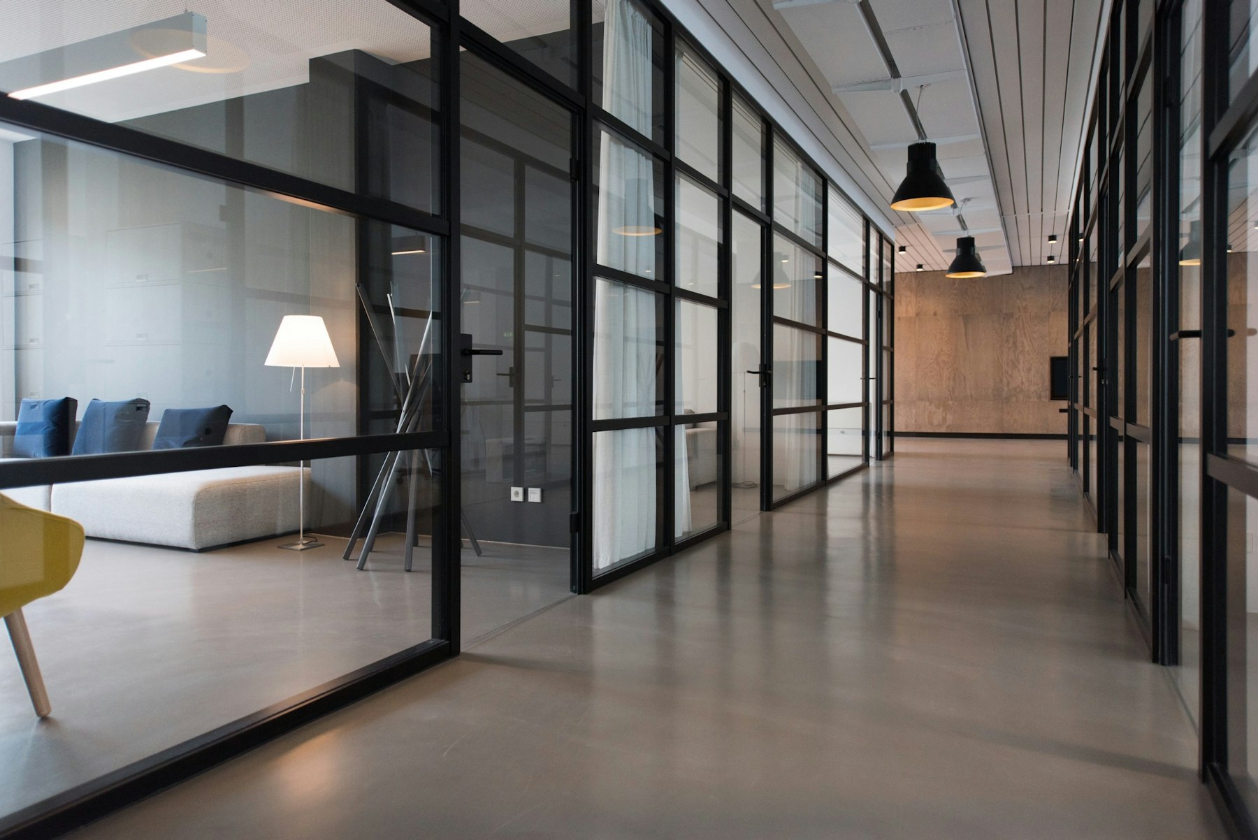 Office corridor lined with steel-framed glass partitions, polished concrete floor and pendant lighting