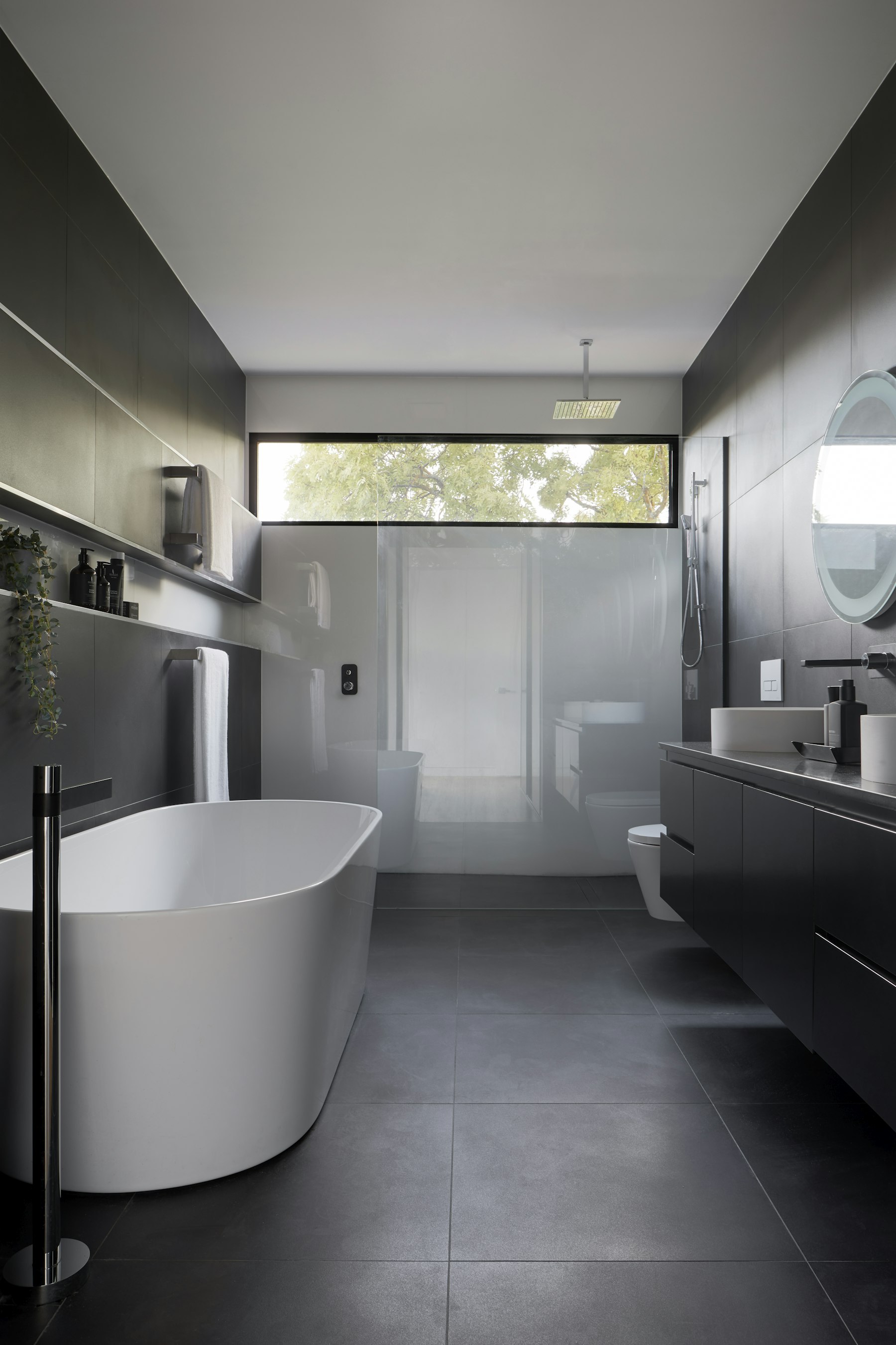 Charcoal-tiled bathroom with a freestanding tub below a long clerestory window and a floating vanity